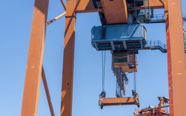 Büyük bir turuncu kargo vinci bir nakliye bölgesinin üzerine yerleştiriliyor, mavi konteynırları parlak mavi gökyüzüne doğru kaldırıyor, limandaki hareketliliği gösteriyor..