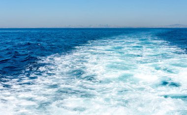 Bir tekne, köpüklü beyaz dalgalar bırakarak, parlak turkuaz suyu kesiyor. Uzak bir şehrin ufuk çizgisi açık mavi bir gökyüzünün altında zar zor görülebilir..