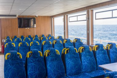 Canlı mavi koltuklar bir feribotun kabinini dolduruyor, berrak bir gökyüzünün altındaki ışıldayan okyanusu net bir şekilde görüyor. Güneş ışığı geniş pencerelerden akar.