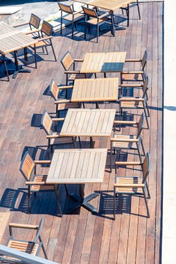 Güneş ışığı, ahşap mobilyaların düzgünce yerleştirildiği rahat bir açık hava yemek alanını yıkıyor. Masalar ve sandalyeler misafirleri bekliyor, sakin sahil atmosferini ve davetkar ortamını vurguluyorlar..