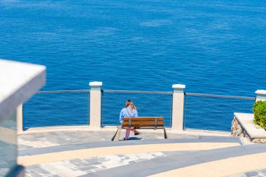 Bir kişi bir bankta sessizce oturur, dingin mavi sulara bakar. Açık gökyüzü konumun sükunetini arttırıyor, yansıma için mükemmel bir kaçış sunuyor..