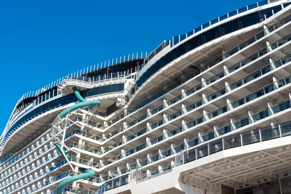 Devasa bir yolcu gemisi, berrak mavi gökyüzünün altında modern mimari tasarımı gösteren canlı bir su kaydırağını ve balkonları sergiliyor. Gemi yanaştı, turistleri gemiye davet ediyor..