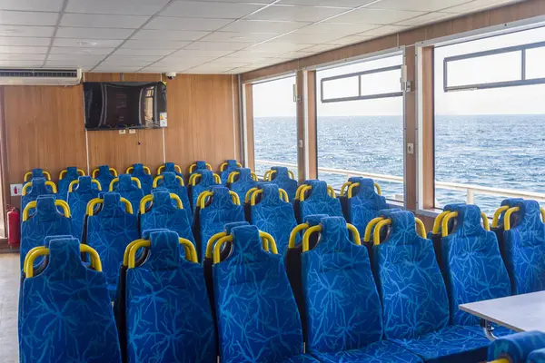 Canlı mavi koltuklar bir feribotun kabinini dolduruyor, berrak bir gökyüzünün altındaki ışıldayan okyanusu net bir şekilde görüyor. Güneş ışığı geniş pencerelerden akar.