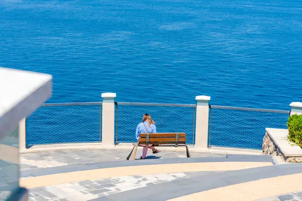 Bir kişi bir bankta sessizce oturur, dingin mavi sulara bakar. Açık gökyüzü konumun sükunetini arttırıyor, yansıma için mükemmel bir kaçış sunuyor..