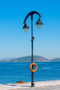 Fenerler limanı aydınlatırken can simidi direkten sarkıyor. Arka planda, tepeler parlak bir gökyüzünün altındaki sakin mavi suların üzerinde yükselir..