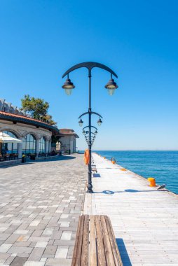 Manzaralı bir gezinti güvertesinde kaldırım boyunca uzanan zarif sokak lambaları var. Parlak güneş ışığı sakin suyu aydınlatır, büyüleyici bir binanın yakınında huzurlu bir atmosfer yaratır..