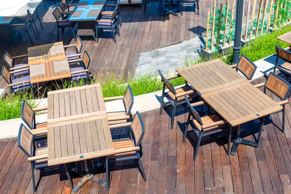 Geniş bir açık hava yemek alanı ahşap döşeme üzerinde ahşap masalar ve sandalyeler düzgünce dizilmiş. Güneş yeşilliklerle dolu bu davetkar atmosferde parlıyor..