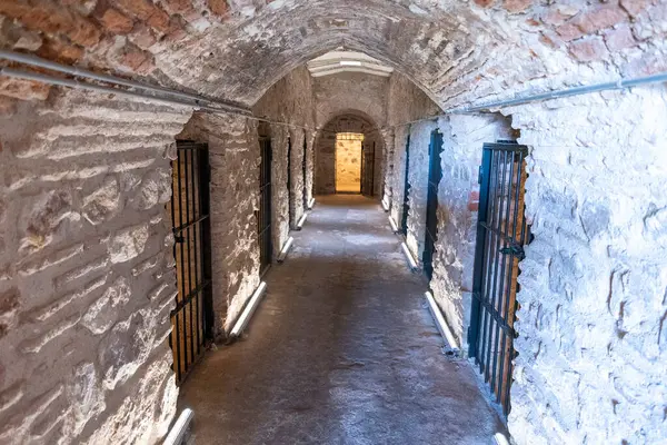 Dar bir koridorda, tarihi bir hapishane ortamını gösteren sert taş duvarlar ve parmaklıklı kapılar yer alıyor. Modern ışıklandırma, geçmiş ve şimdiki zaman arasında benzersiz bir zıtlık oluşturarak hatları vurgular..