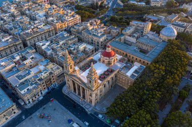 Malta 'daki Floriana Parish Kilisesi' nin havadan görünüşü, büyük Barok cephesini, ikiz çan kulelerini ve çarpıcı kırmızı kubbesini gözler önüne seriyor. Yoğun kentsel mimari ortamına kurulmuş ve gün batımında ağaçlarla çevrili yemyeşil yollarla çevrili..