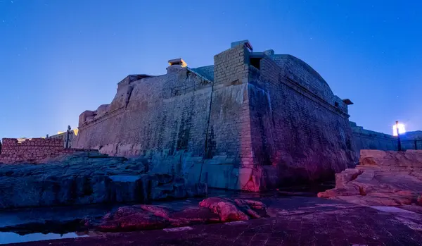 Valletta Malta 'daki St. Elmo Kalesi geceleri aydınlatılır. Elmo Valletta, Malta 'da, geceleri aydınlanıyor. Tarihi kalenin devasa taş duvarları, gece gökyüzü ile zıt mavi ve mor renklerle kaplıdır..