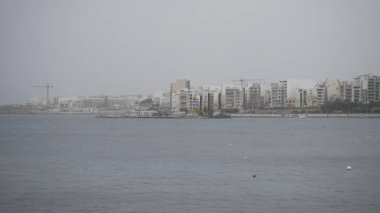 Malta 'da Bugibbas deniz cephesi bulutlu bir gökyüzünün altında, sakin sulara bakan apartmanların kentsel ufuk çizgisi.