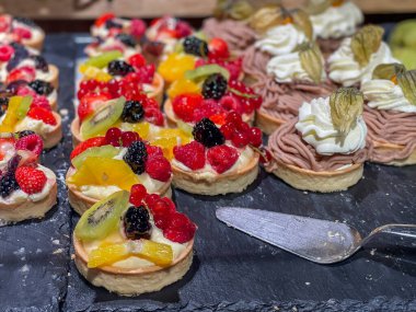 Pasta çeşitleri. Renkli böğürtlenli ve meyveli mini meyveli pasta ve kremalı kestane ezmesi, İsviçre yerel çöl vermicellleri, siyah taş tepside sergileniyor.
