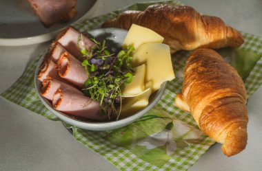 Sabah kahvaltısı. jambon, peynir ve lahanalar kasede, taze kruvasanlar yeşil örtüde, güneş ışınları ve yüksek açılı gölgeler.