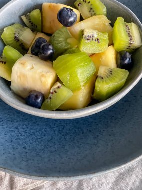 Taze tropik meyve salata çölü mavi seramik kasede çömlek tabağı ve keten peçete, renkli karışık ananas, kivi, yabanmersini, yüksek açılı manzara, sağlıklı yaşam tarzı konsepti.