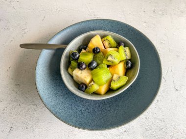 Taze tropikal meyve salatalı tatlı mavi seramik kasede kil tabakta beyaz taş mermer zemin üzerinde, renkli karışım ananas, kivi, yabanmersini, yüksek açı görünümü, kopyalama alanı