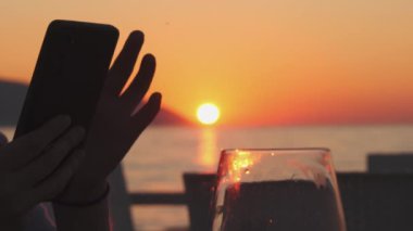 Smartphone 'daki kız WIne içiyor ve Seaside Sunset' in yanında oturuyor. Yüksek kalite 4k görüntü