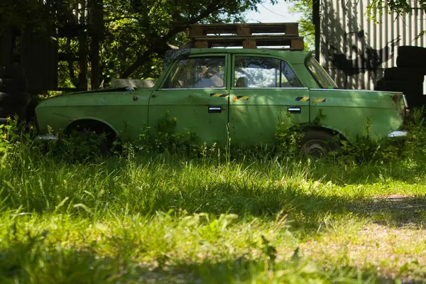 stock image old green car abandoned surrounded by abandoned buildings overgrown with bright green grass