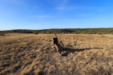 Bir kız, sarı çimenli ve mavi gökyüzünün altında büyük bir taşın üzerinde oturuyor.