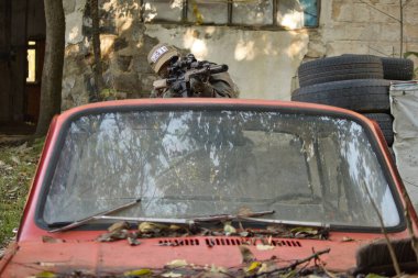 Askeri teçhizatlı bir kişinin eski bir arabanın koruduğu bir tüfeği hedef aldığı bir taktik sahnesi. Arka planda lastikler ve yıpranmış duvarlar, eğitim sahası veya savaş senaryosu atmosferi yaratıyor.