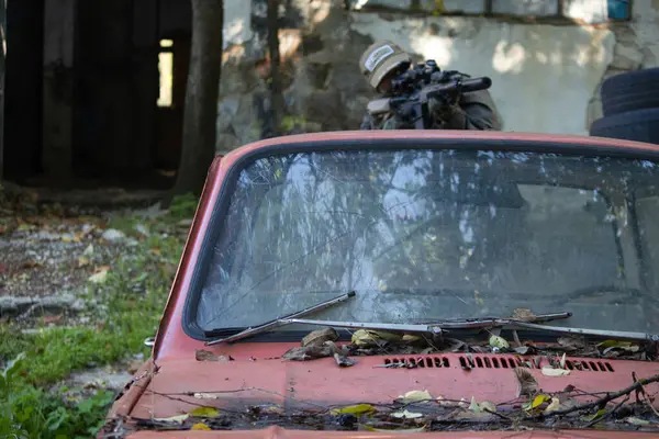 Askeri teçhizatlı bir kişinin eski bir arabanın koruduğu bir tüfeği hedef aldığı bir taktik sahnesi. Arka planda lastikler ve yıpranmış duvarlar, eğitim sahası veya savaş senaryosu atmosferi yaratıyor.
