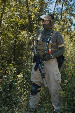 A man in tactical gear prepares his airsoft gun during an outdoor game. Wearing protective goggles, gloves, and a vest, he focuses on safety and precision in the field.