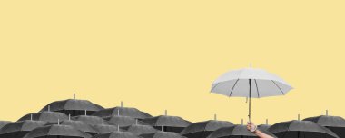 Açık sarı arka planda diğer siyah şemsiyelerin üzerinde beyaz bir şemsiye tutuyor..