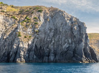 Yıpranmış deniz kenarı kaya yüzeyi dokusu ile yeşil ve mavi su parçaları. Yaşlanmış volkanik duvar yüzeyinin arka planında çatlaklar ve çizikler var. Ischia Adası, İtalya.