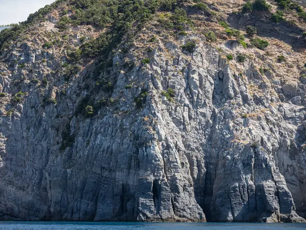 Yıpranmış deniz kenarı kaya yüzeyi dokusu ile yeşil ve mavi su parçaları. Yaşlanmış volkanik duvar yüzeyinin arka planında çatlaklar ve çizikler var. Ischia Adası, İtalya.