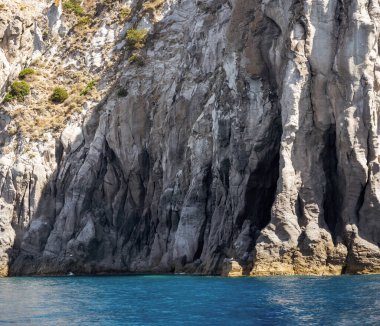 Yıpranmış deniz kenarı kaya yüzeyi dokusu ile yeşil ve mavi su parçaları. Yaşlanmış volkanik duvar yüzeyinin arka planında çatlaklar ve çizikler var. Ischia Adası, İtalya.