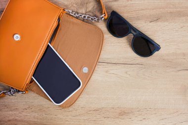 İçinde akıllı telefon ve ahşap arka planda güneş gözlüğü olan turuncu kadın vücut çantası..