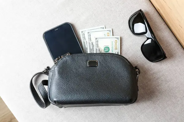 stock image Black leather crossbody bag with smartphone and dollars on the pouf.