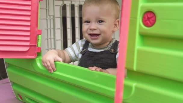 幸せな笑顔の赤ちゃん男の子子供はかくれんぼをしてプラスチック製の遊び場の窓を見ています 庭の外で小さなアクティブな子供の遊び遊び場 — ストック動画