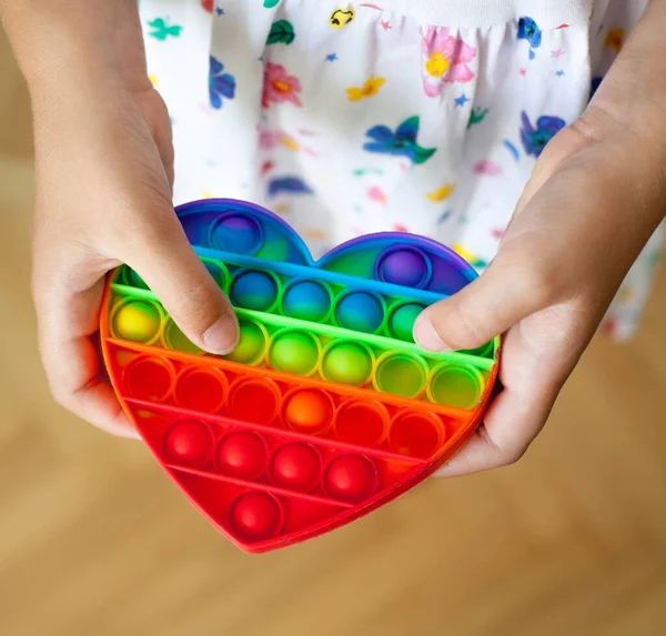 ハート型の虹を持った女の子がおもちゃをポップします 新しいフィジェットトレンディーなおもちゃ ストレス対策のおもちゃ シンプルなディンプル — ストック写真