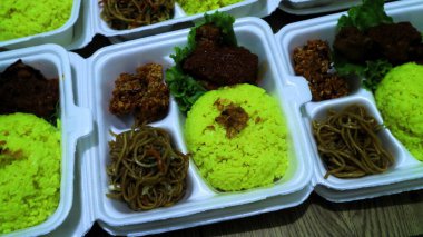 Büyük miktarda sarı pirinç, erişte ve köpüklü yiyecek kabında servis edilen ızgara tavuk paketleri.