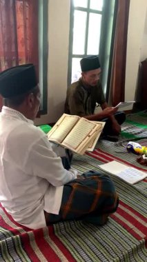 Asyalı Endonezyalı erkekler sarong ve peci giyip oturma odasında Kur 'an okuyorlar, Müslümanlar için dini günlük keskinlik.