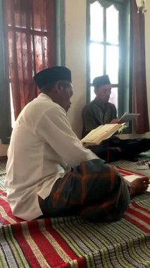 Asyalı Endonezyalı erkekler sarong ve peci giyip oturma odasında Kur 'an okuyorlar, Müslümanlar için dini günlük keskinlik.