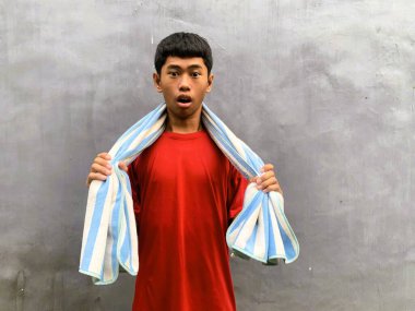A surprised and startled southeast asian teenager in a red t-shirt with a towel on their shoulder, against a plain gray background