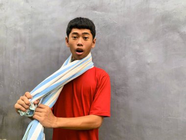 A surprised and startled southeast asian teenager in a red t-shirt with a towel on their shoulder, against a plain gray background