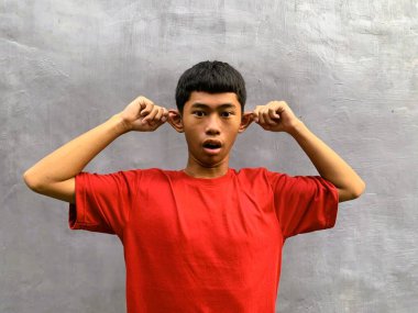 A surprised, startled, and funny southeast asian teenager pulls their own ear with an expressive face