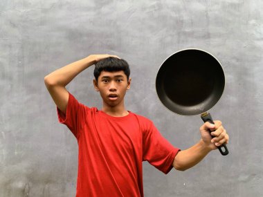 A southeast asian teenager with shocked and startled expression in a red t-shirt carries a frying pan