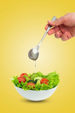 Beyaz salatalı salata, salatalık, domates, buzdağı salatası yaprakları ve erkek eli sarı arka planda izole edilmiş salatanın üzerine kaşıkla yağ döküyor.