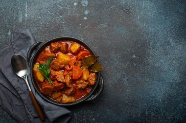 Patatesli sığır eti yahnisi, havuç ve siyah güveç tenceresinde leziz sos defne yapraklarıyla ve yukarıdan gelen koyu kırsal beton arka planda kaşıkla taze yeşil otlar, mesaj için yer..