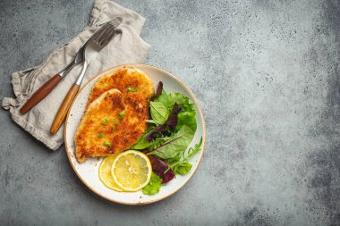 Crispy panko breaded fried chicken fillet with green salad and lemon on plate on gray rustic concrete background table from above. Japanese style deep fried coated chicken breasts, space for text.