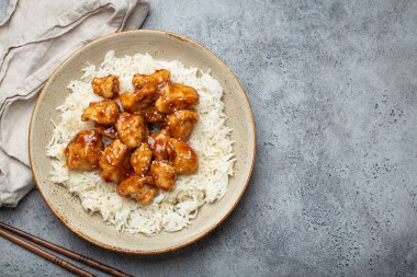 Chinese sweet and sour sticky chicken with sesame seeds and rice on ceramic plate with chopsticks top view on gray rustic stone background, traditional dish of China. Space for text