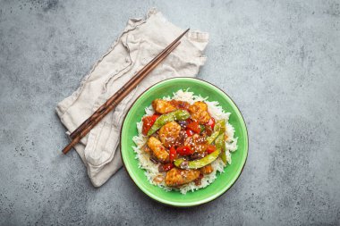 Asian sweet and sour sticky chicken with vegetables stir-fry and rice in ceramic bowl with chopsticks top view on gray rustic stone background, traditional Asian dish
