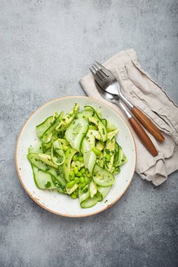 Healthy vegan green avocado salad bowl with sliced cucumbers, edamame beans, olive oil and herbs on ceramic plate top view on grey stone rustic table background