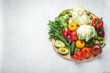 Rustic wooden tray with selection of fresh vegetables and greenery on white kitchen table top view. Vegetarian or diet food from organic ingredients, healthy nutrition concept, space for text