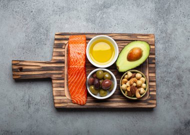 Food sources of healthy unsaturated fat: fresh raw salmon fillet, avocado, olives, nuts on cutting board on rustic stone background top view. Omega 3 and oil products, healthy nutrition and keto diet