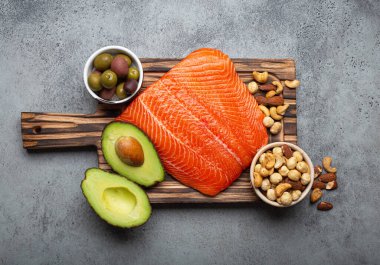 Food sources of healthy unsaturated fat: fresh raw salmon fillet, avocado, olives, nuts on cutting board on rustic stone background top view. Omega 3 and oil products, healthy nutrition and keto diet
