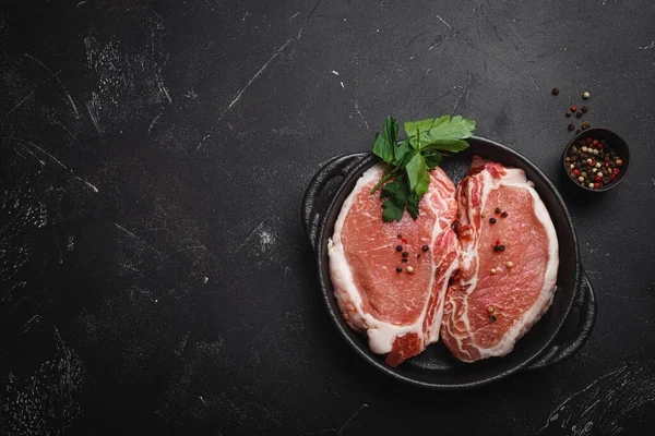 Cut raw meat pork steaks with seasonings in black cast iron pan, dark rustic stone background top view, ready for roasting. Pork loin chops cooking with space for text